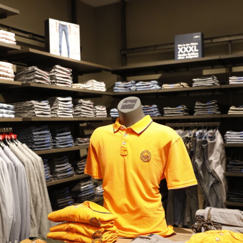 Ausgestelltes gelbes Polo-Shirt mit Verkaufsregalen im Hintergrund in der engbers-Filiale im Forum Köpenick.